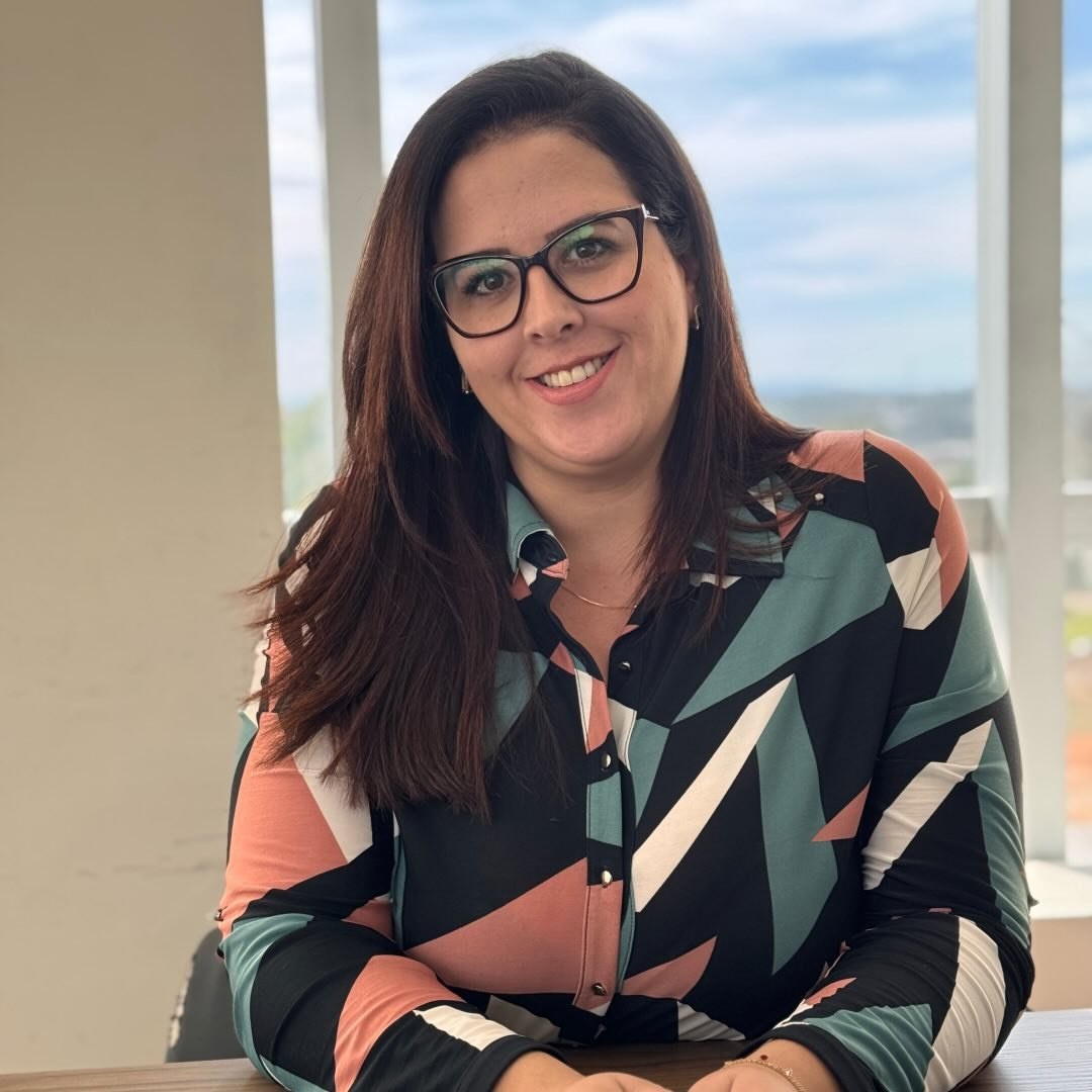 Renata Pinto, advogada previdenciarista, sorrindo, sentada à mesa diante de uma janela